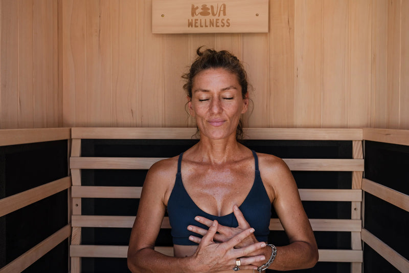 A woman sits with her eyes closed and hands on her chest beneath a Kiva Wellness sign inside a sauna.