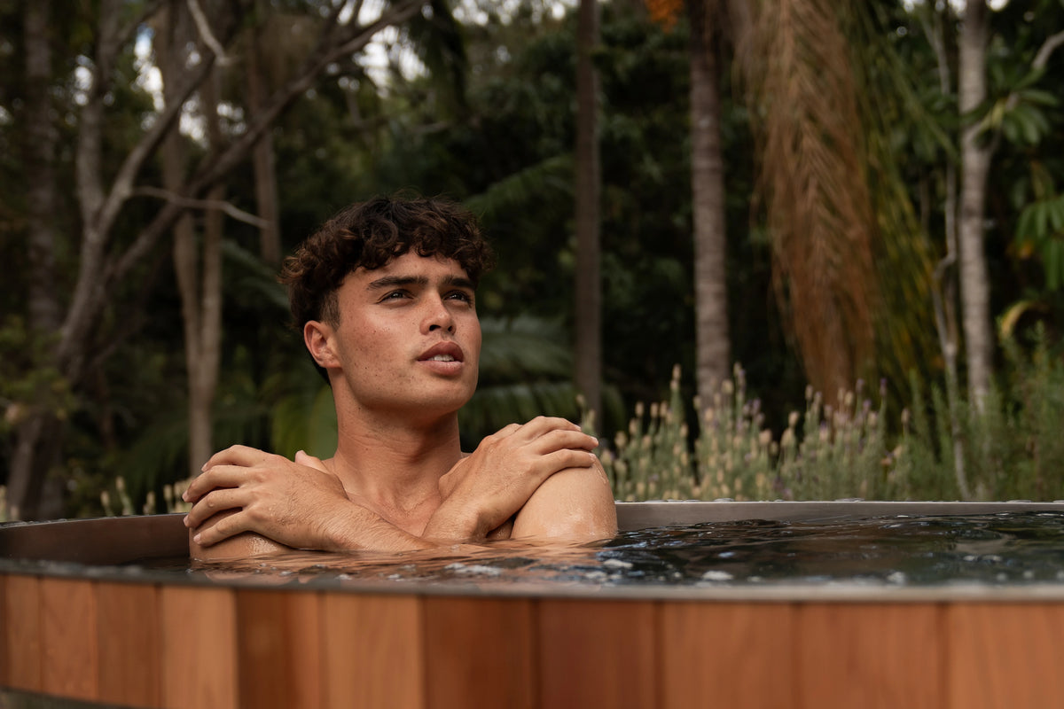 A young man rests his crossed arms on the rim of a cedar ice bath in a leafy garden.