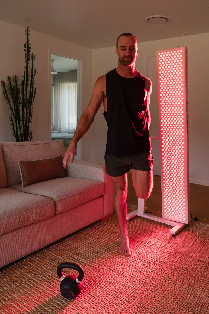 A man stretches one leg while standing beside a red light therapy panel, with a kettlebell on the floor.
