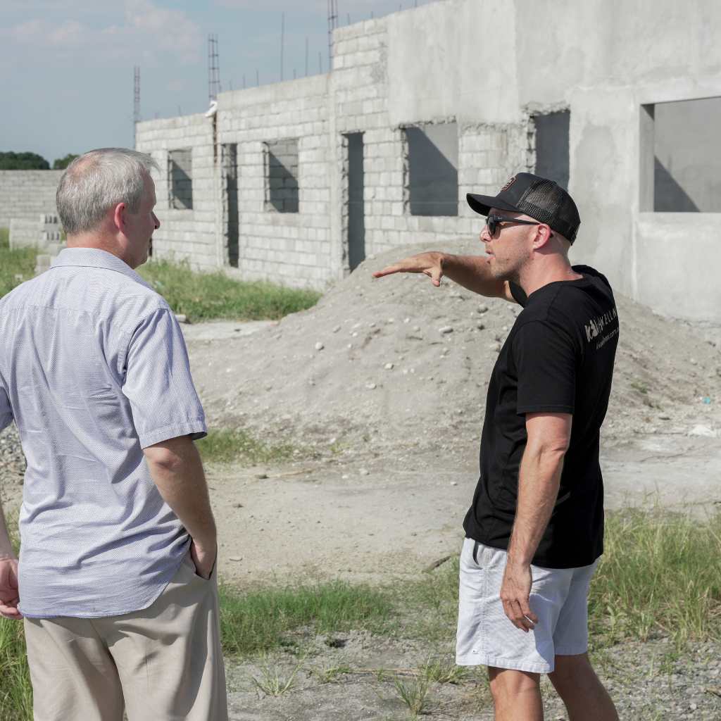 Two men inspecting a partly built concrete block building with one pointing towards the structure.