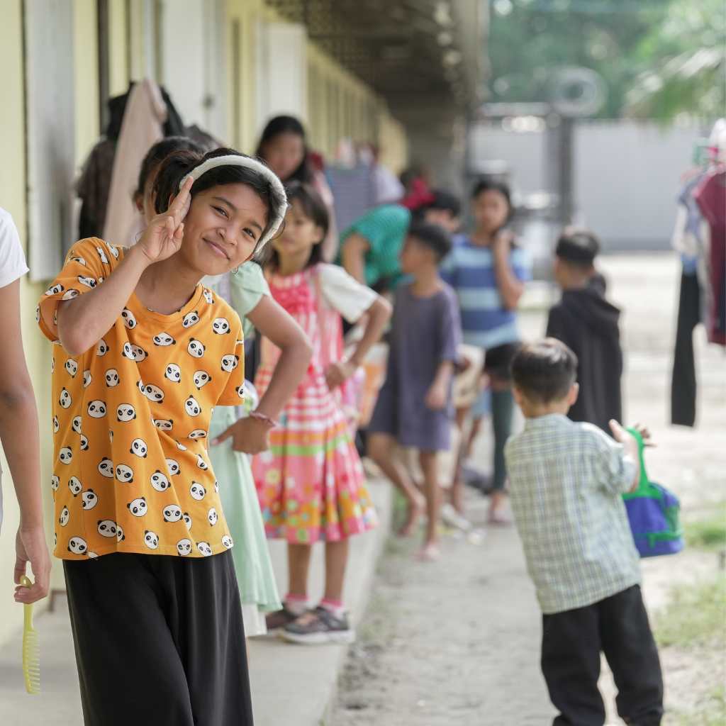 Children lined up outside a yellow classroom building with a girl waving at the front.