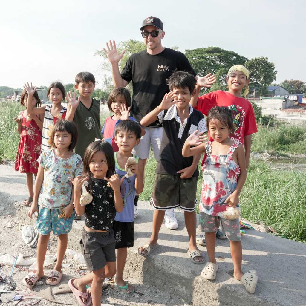 Man in a black cap standing among a group of children waving outdoors.