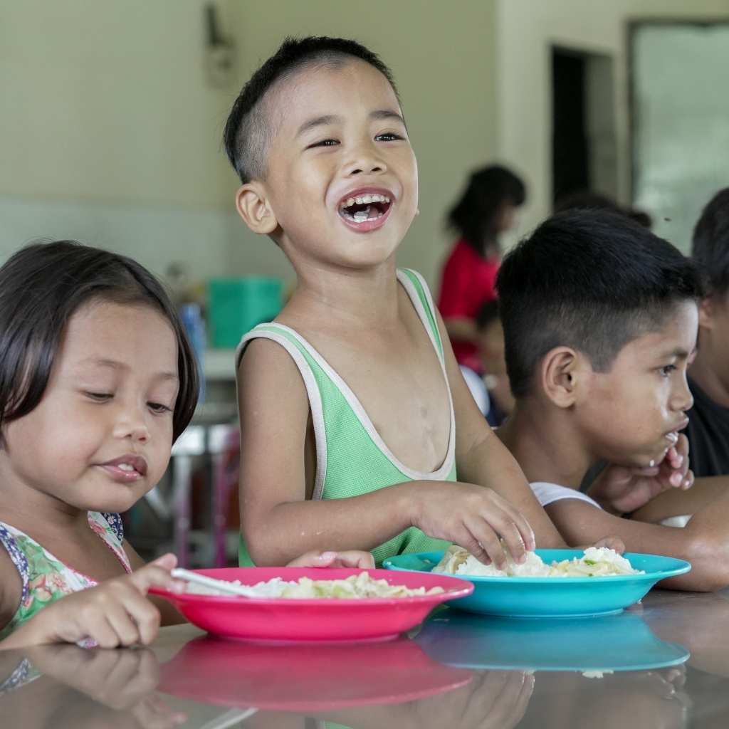 Smiling boy seated beside other children at a long table with plates of food.