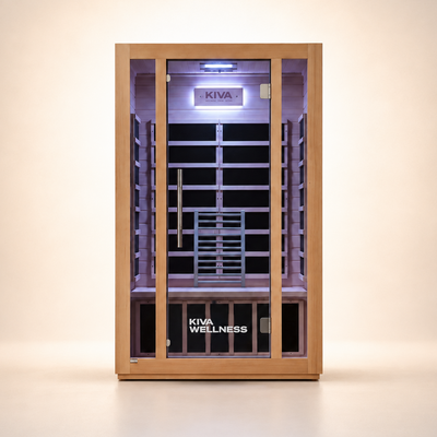 A Kiva Wellness infrared sauna with a light natural wood exterior, glass door, and a dark interior with wooden benches and a backrest, shown on a clean beige studio background.