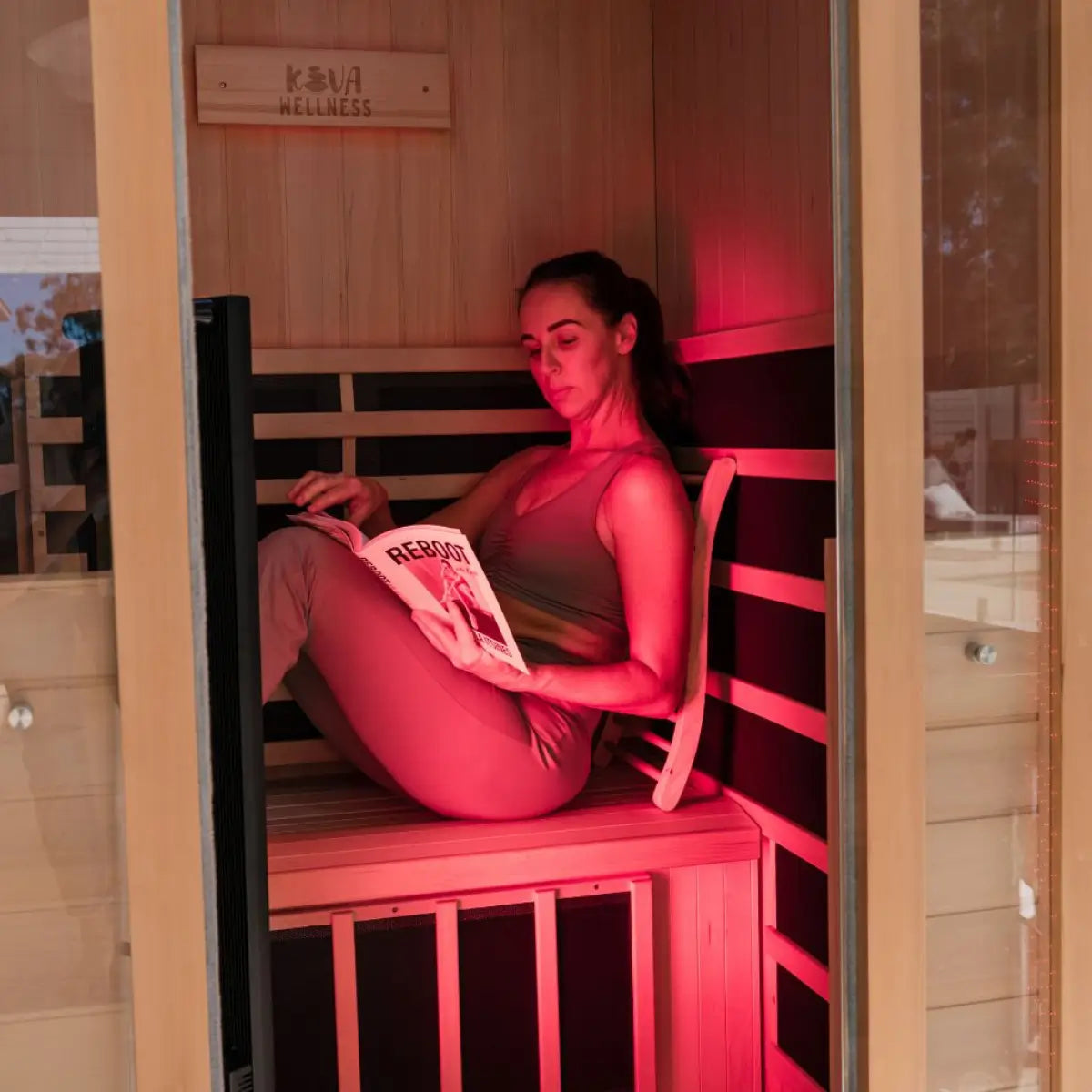 A woman reads on a timber sauna bench beside an illuminated Kiva Wellness Radiance red light therapy panel.