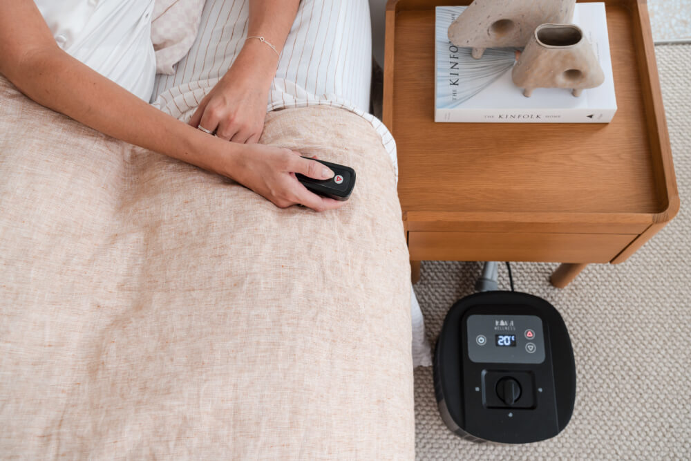 Person in bed holding a remote control with a sleep cooling unit on the floor beside the bedside table.