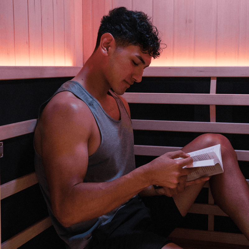 Man reading a book on a timber sauna bench under warm red lighting.