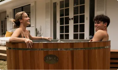 A man and woman smile at each other from opposite sides of a round cedar ice bath outside a house.