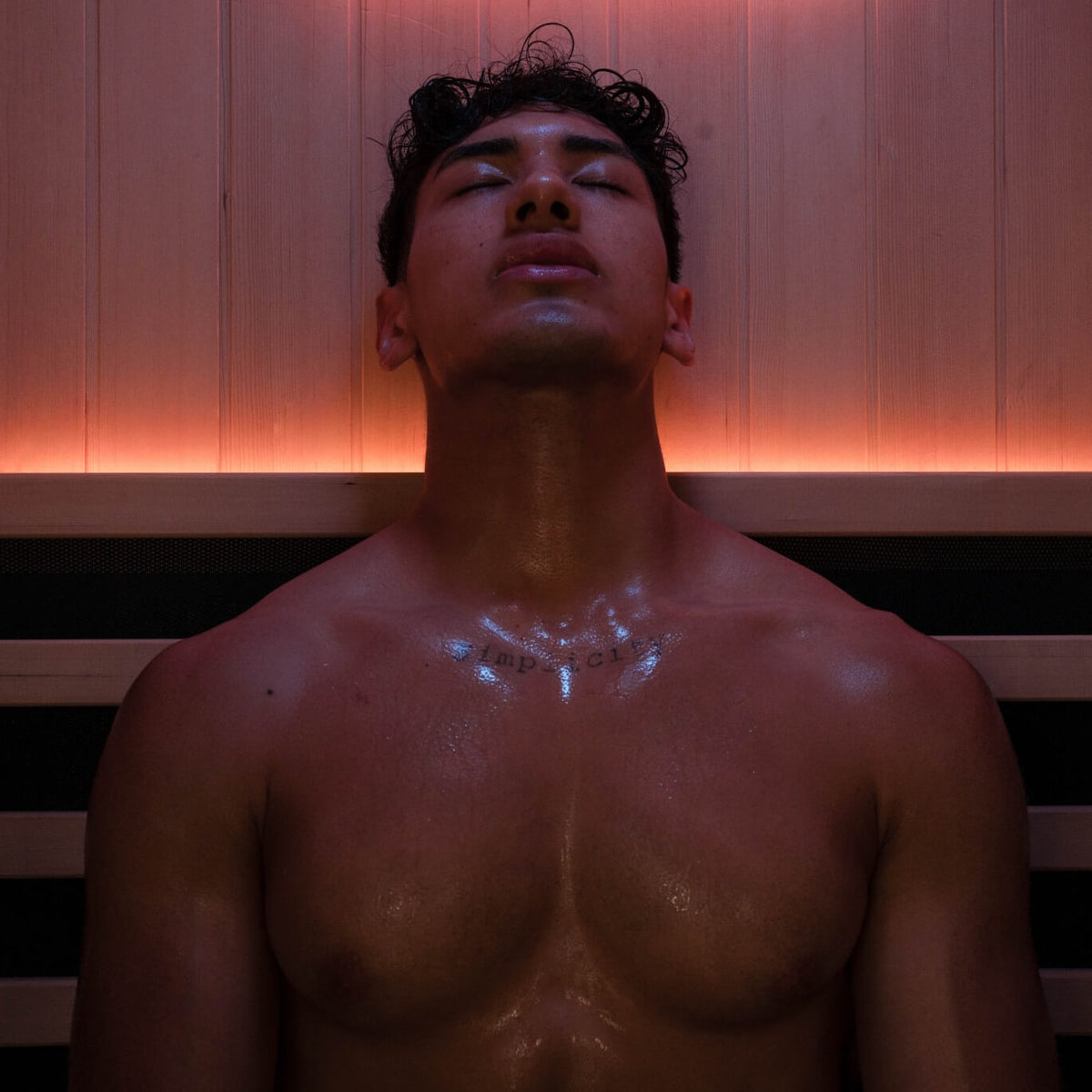 Man sitting with his eyes closed and head tilted back under warm red sauna lighting.