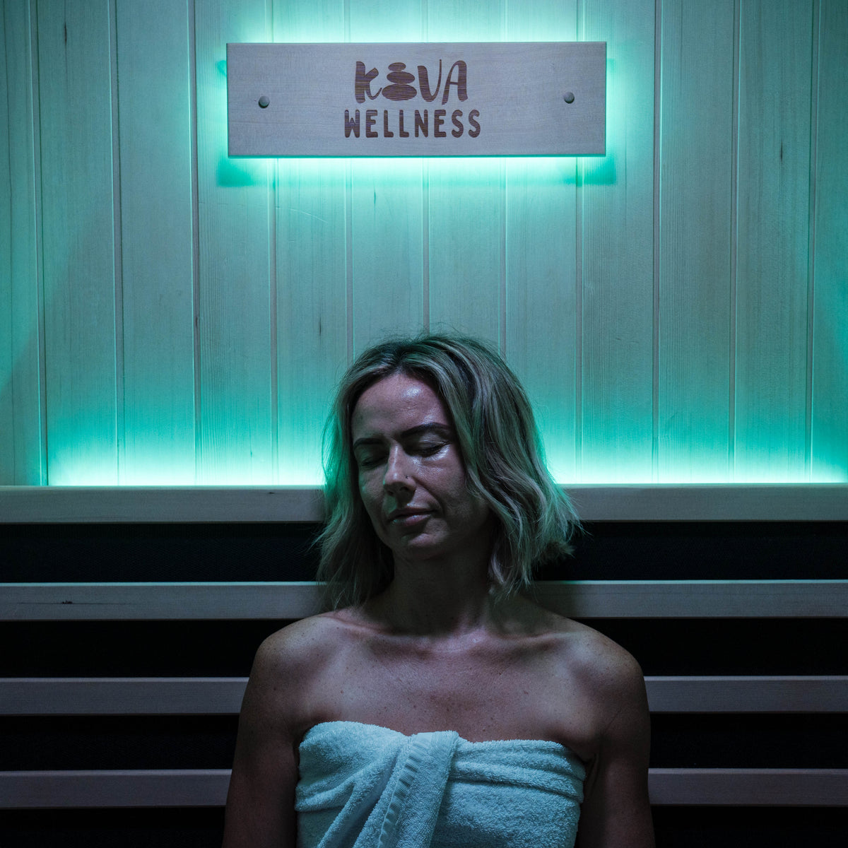 Woman wrapped in a towel resting beneath the Kiva Wellness sign under blue sauna lighting.