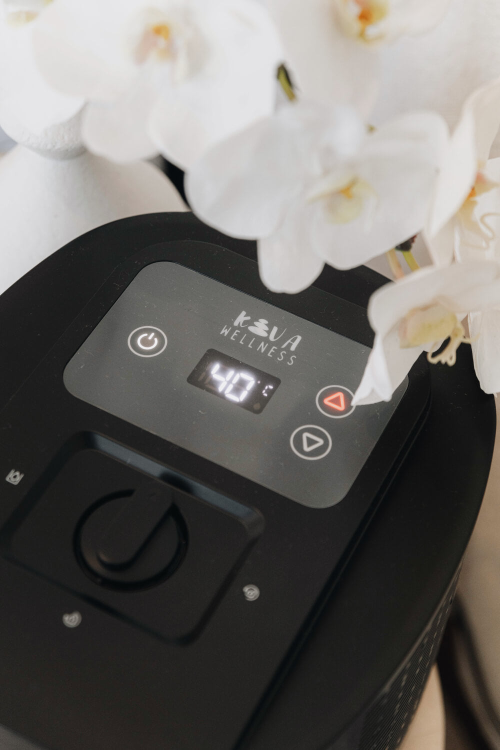 Close view of a black Kiva Wellness sleep cooling unit's digital controls beneath white orchid flowers.
