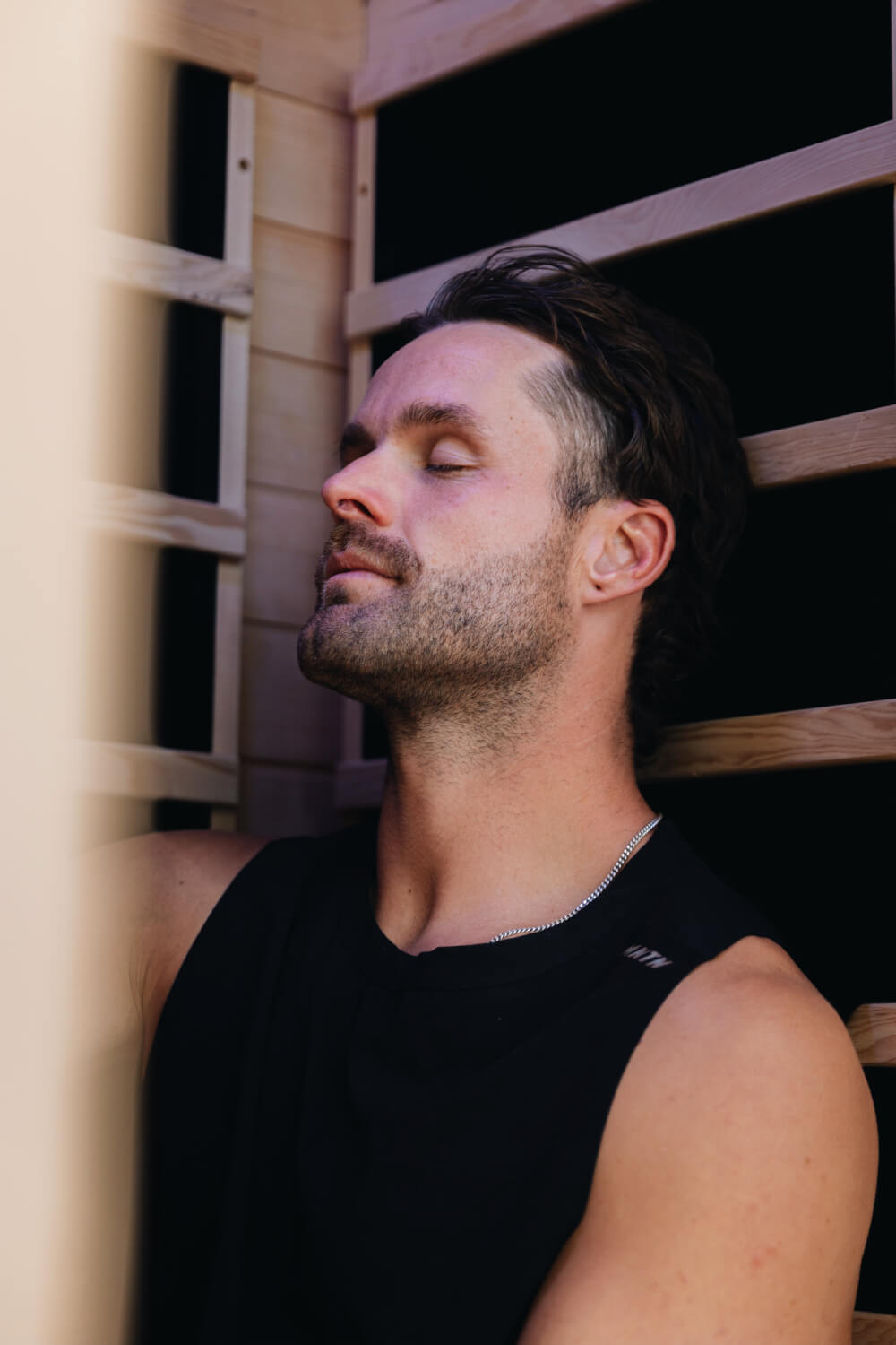 Man in a black sleeveless top resting with his eyes closed against a timber sauna backrest.