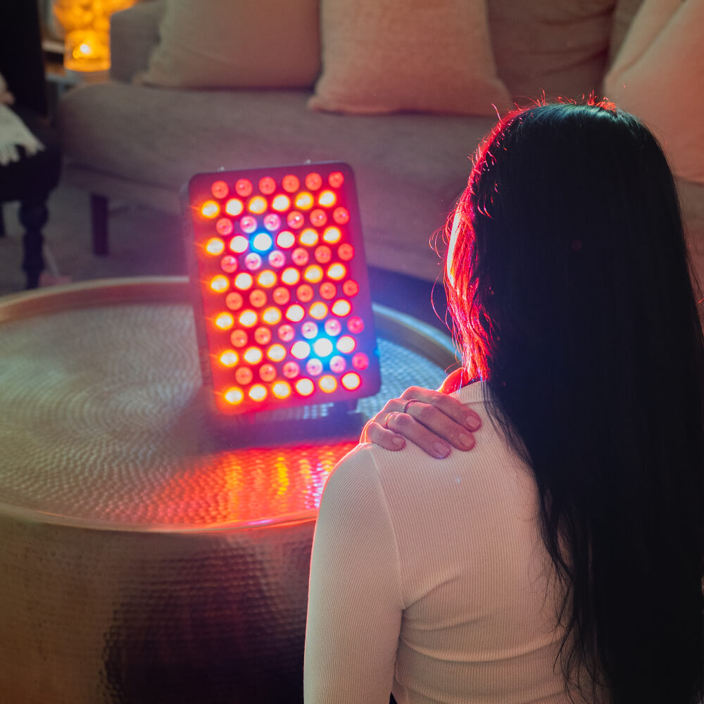 Woman seated facing an illuminated Solace Glow red light therapy panel on a living room table.