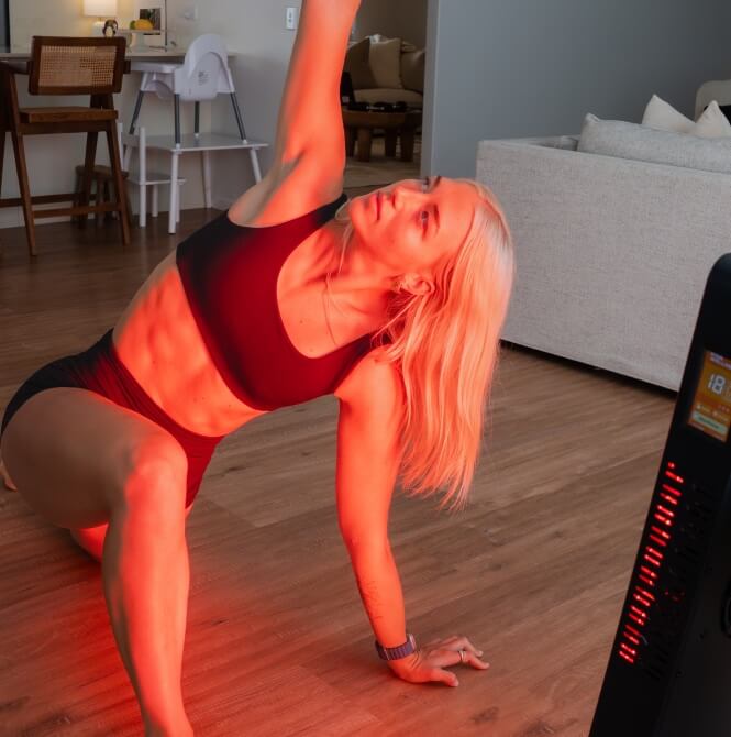 Woman performing a side stretch on the floor beside an illuminated red light therapy panel.