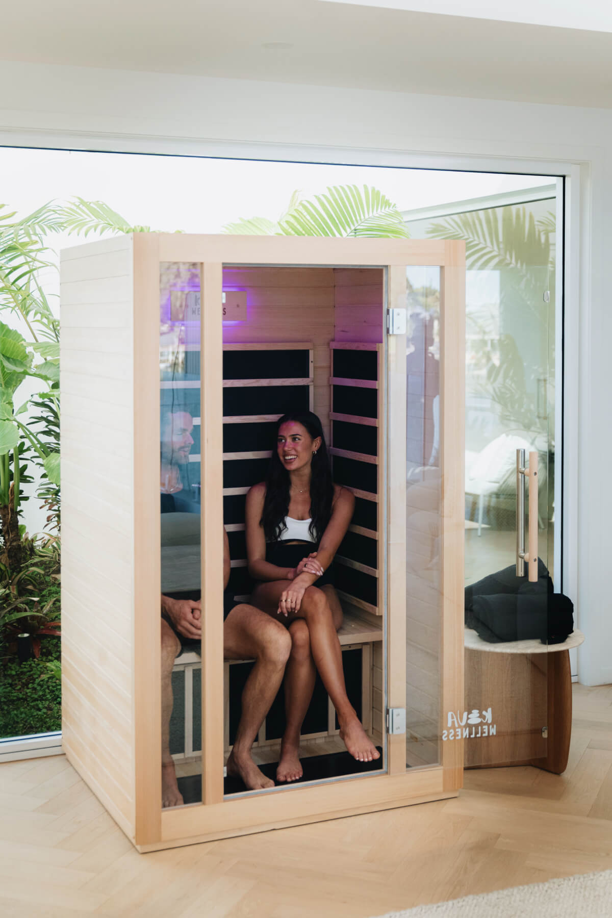 Two women seated inside a Kiva Wellness Rise sauna beside large windows and potted greenery.