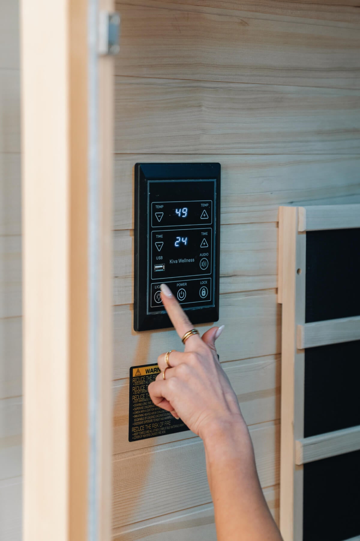Hand pressing the digital control panel on a pale timber sauna wall beside a heater panel.