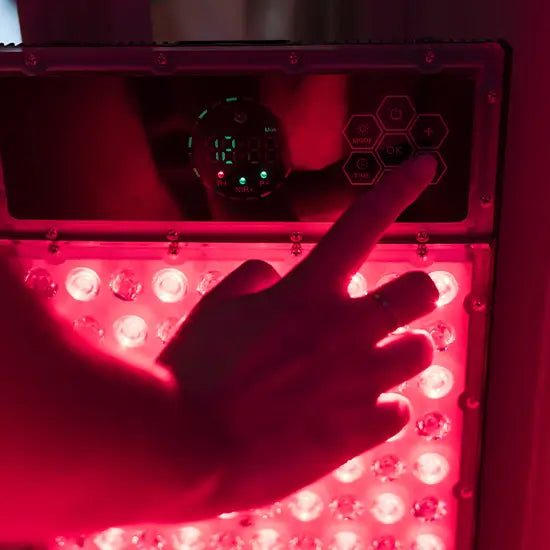 A hand adjusts the control buttons above the glowing LEDs on a Kiva Wellness Radiance sauna red light panel.