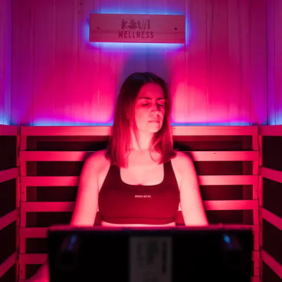 A woman relaxes with her eyes closed behind a Kiva Wellness Radiance red light panel inside a timber sauna.