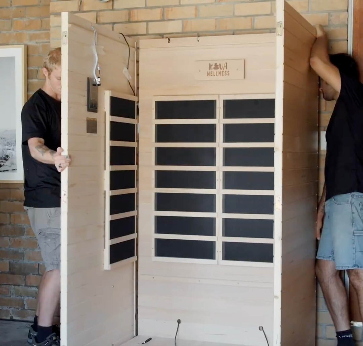 Two people assembling a timber sauna cabin around its upright wall panels.
