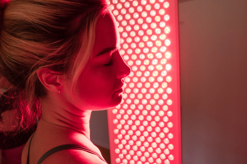 Profile of a woman standing close to a red light therapy panel with its LEDs illuminating her face.