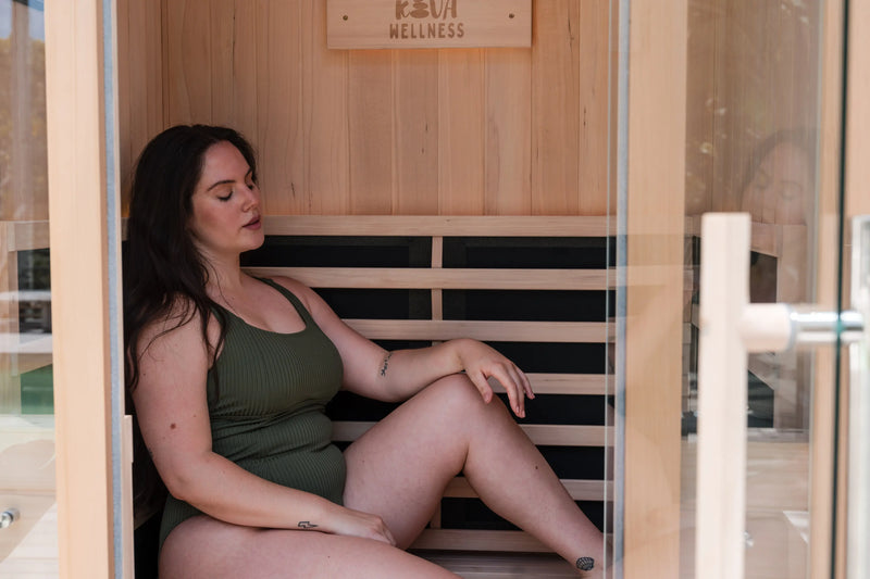A woman in a green swimsuit relaxes on the bench inside a Kiva Wellness sauna with the glass door open.