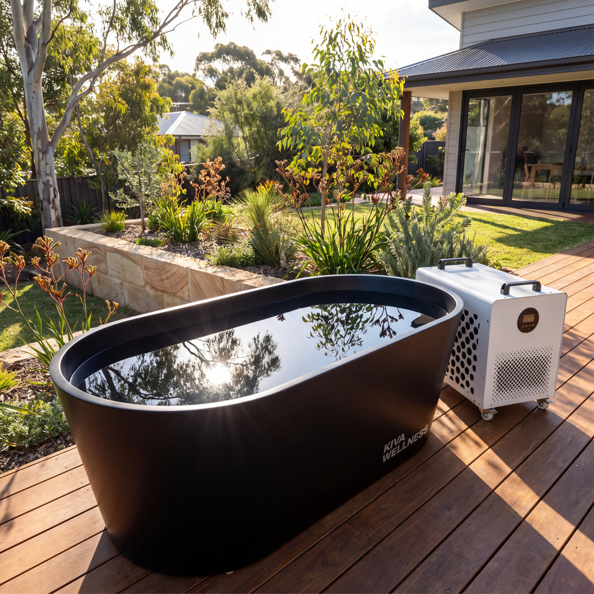 Kiva Kaldr black ice bath filled with water placed on a wooden deck beside  the Core chiller in a sunny backyard garden setting.