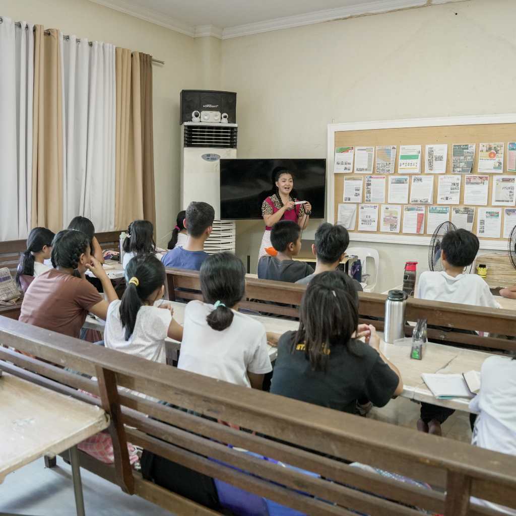 Children seated at benches in a classroom facing a teacher at the front.