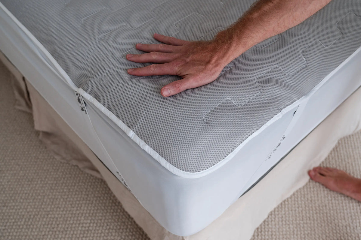 A hand presses down on the grey quilted surface of a mattress topper fitted to a bed.