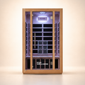 A Kiva Wellness infrared sauna with a light natural wood exterior, glass door, and a dark interior with wooden benches and a backrest, shown on a clean beige studio background.