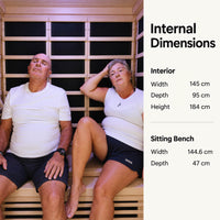 A man and a woman sit inside a wooden 3 person kiva wellness sauna with text on the right listing internal dimensions: interior width 145 cm, depth 95 cm, height 184 cm, and sitting bench width 144.6 cm and depth 47 cm.