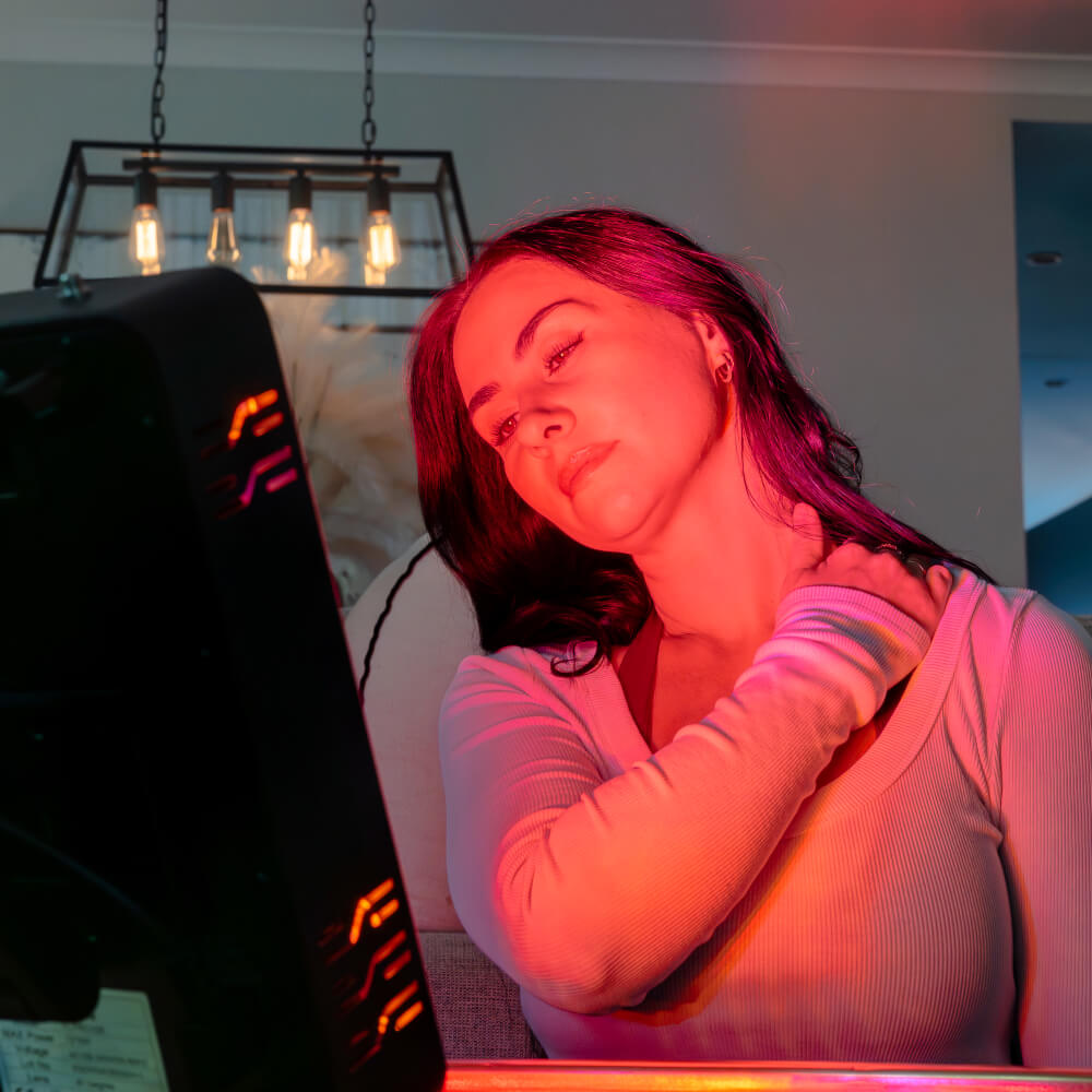 Woman resting a hand on her shoulder while sitting beside a red light therapy panel.