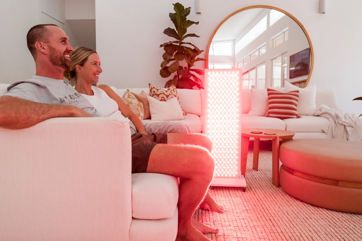A couple sits together on a sofa beside an illuminated red light therapy panel in a bright living room.