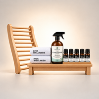 Sauna essentials gift pack arranged on a small wooden tray, including a glass cleaner, 5 essential oils, and two folded towels. The ergonomic sauna backrest is positioned behind the set against a beige background.