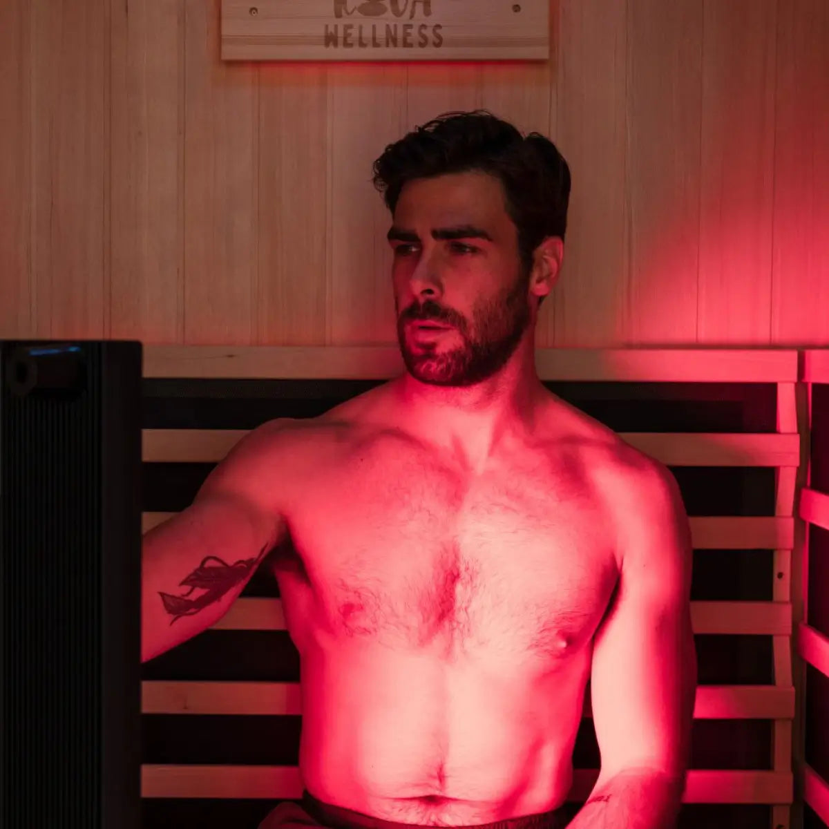 A man sits on a timber sauna bench facing the glow of a Kiva Wellness Radiance red light therapy panel.
