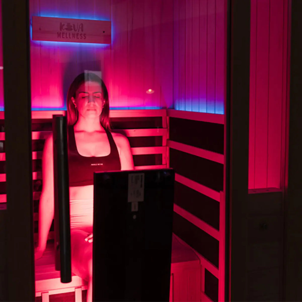 A woman sits inside a timber sauna facing an illuminated Kiva Wellness Radiance red light therapy panel.