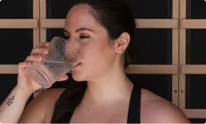 A woman drinks from a clear glass while seated against the slatted wall inside an infrared sauna.