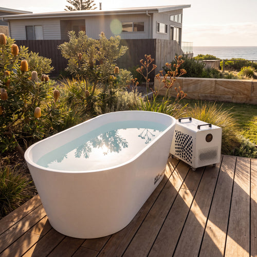Kiva Kaldr white ice bath filled with water sits on a wooden deck outdoors beside the Core chiller, with garden plants, a house, and ocean views in the background.